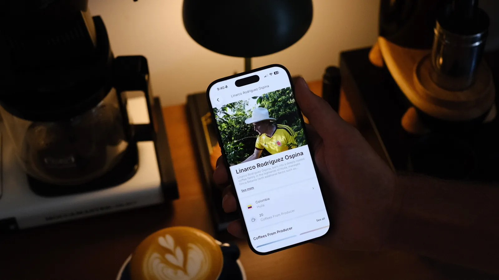 Holding a phone that shows a coffee producer's page in the Siip app, next to a coffee maker and a latte